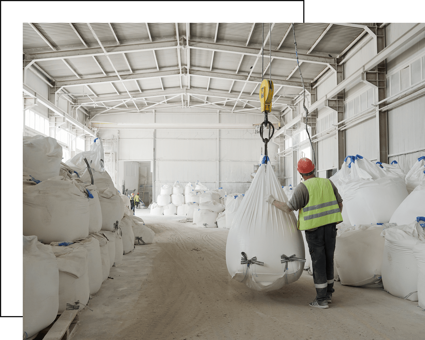 A worker in a high-visibility vest and hard hat expertly guides a large, white industrial storage bag being moved by a crane inside a spacious warehouse filled with similar bags.